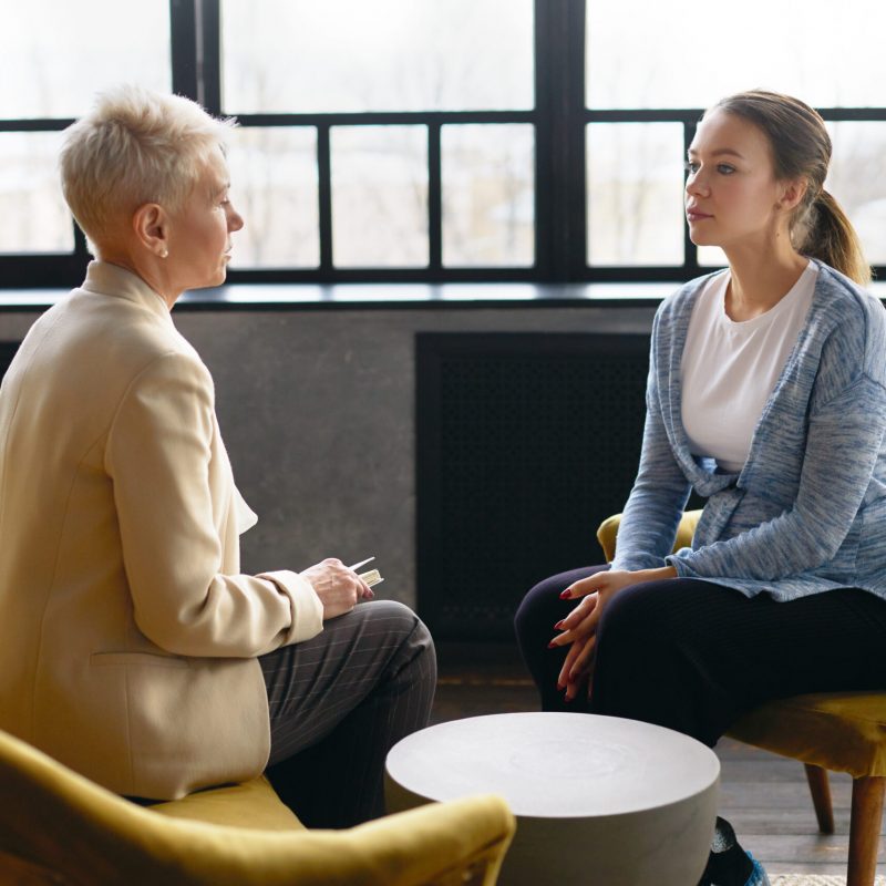 Side view image of two women sitting face to face in comfortable armchairs. Stylish mature female psychotherapist conducting counselling with young pregnant woman. Job interview and human resources